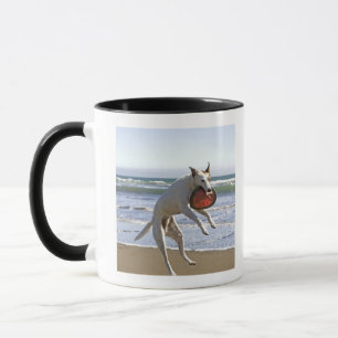 Een hond springt naar een frisbee op het strand mok