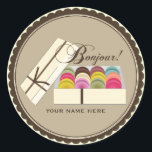 Sticker Rond Une douzaine de Macarons Français Bonjour Personna<br><div class="desc">Un autocollant avec l'illustration d'une douzaine de macarons français colorés dans une boîte cadeau beige avec un ruban brun délicat. Le texte en haut dit "Bonjour !" Personnalisez avec votre nom sous illustration.</div>