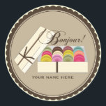Sticker Rond Une douzaine de Macarons Français Bonjour Personna<br><div class="desc">Un autocollant avec l'illustration d'une douzaine de macarons français colorés dans une boîte cadeau beige avec un ruban brun délicat. Le texte en haut dit "Bonjour !" Personnalisez avec votre nom sous illustration.</div>